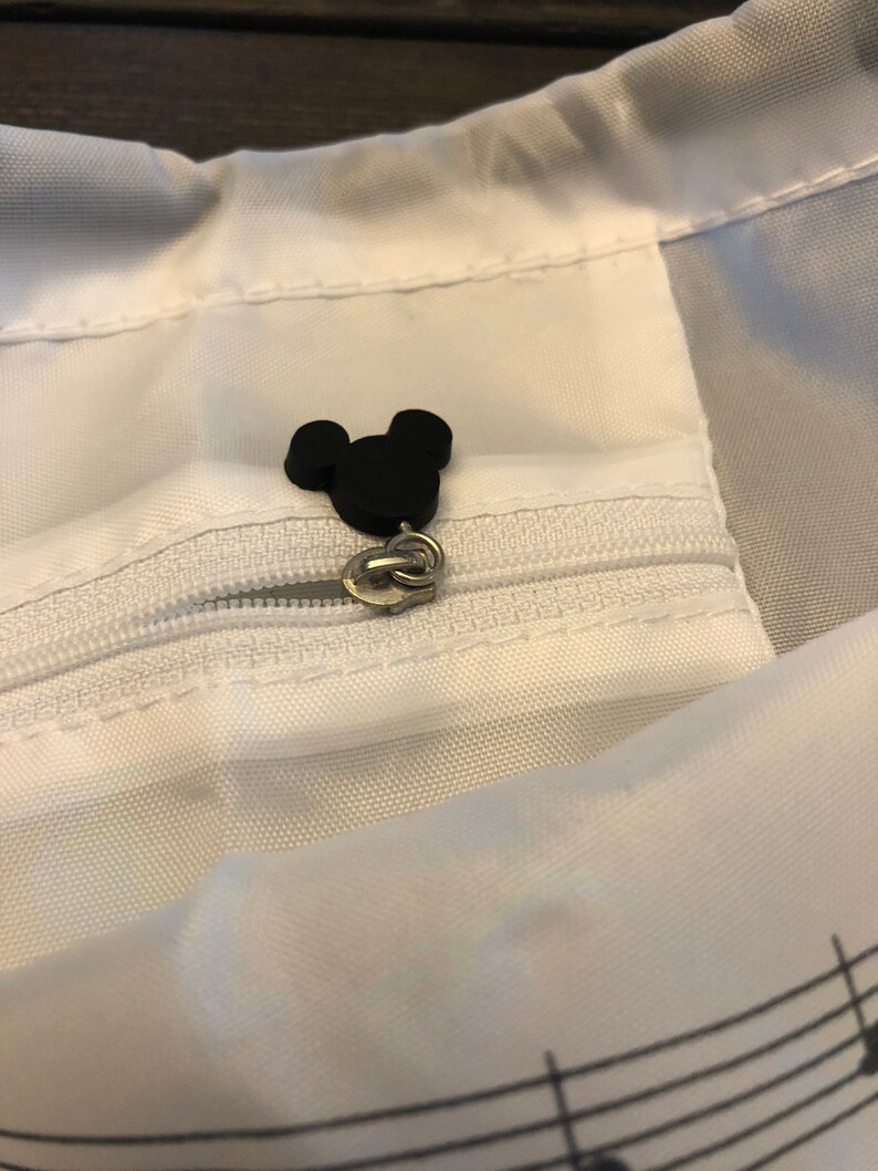 May include: A white drawstring bag with a black Mickey Mouse head zipper pull. The bag has a musical note pattern on the bottom.