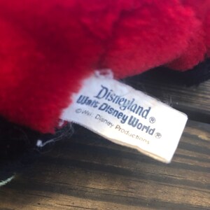 May include: White tag with blue text that reads "Disneyland Walt Disney World" with a copyright symbol and the text "c Walt Disney Productions" with a registered trademark symbol.