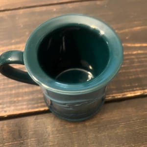 May include: A teal-colored mug with a handle. The mug has a wide rim and a cylindrical body with a decorative embossed pattern. The mug is sitting on a wooden surface.