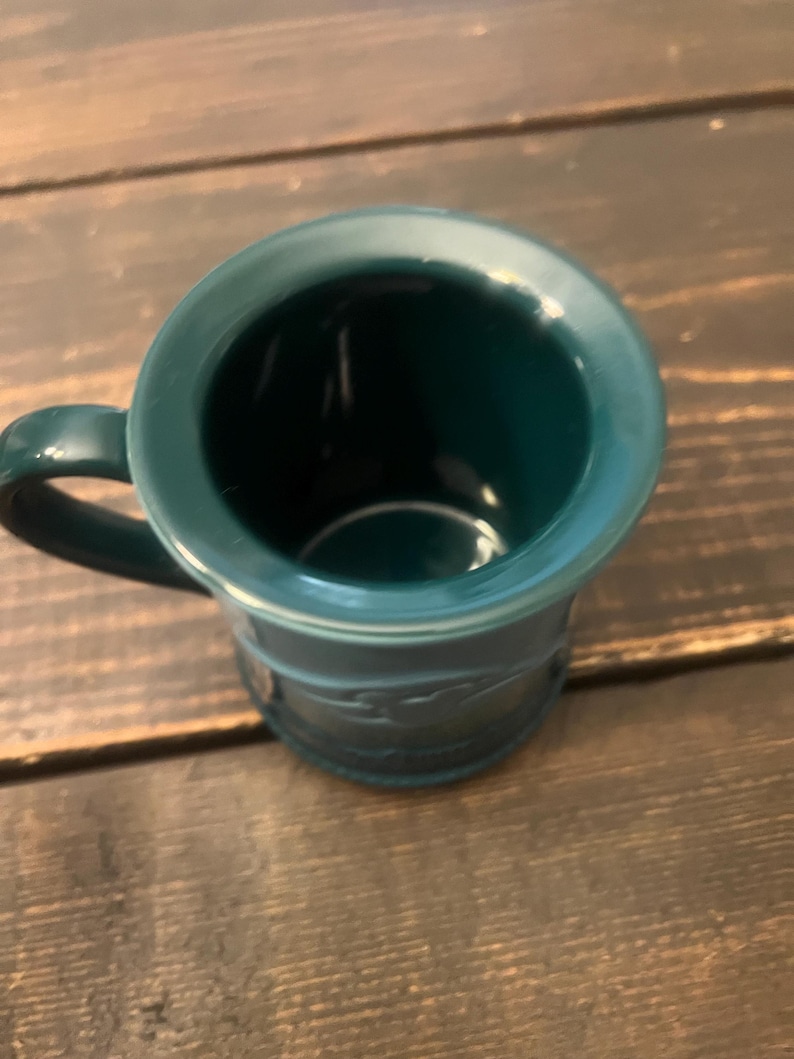 May include: A teal-colored mug with a handle. The mug has a wide rim and a cylindrical body with a decorative embossed pattern. The mug is sitting on a wooden surface.