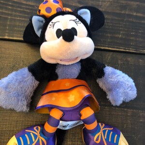 May include: A plush Minnie Mouse doll in a Halloween costume. The doll has a black head and ears, a light pink face, and a bow with orange polka dots. She wears a purple and orange skirt and shoes with orange stripes and bows.