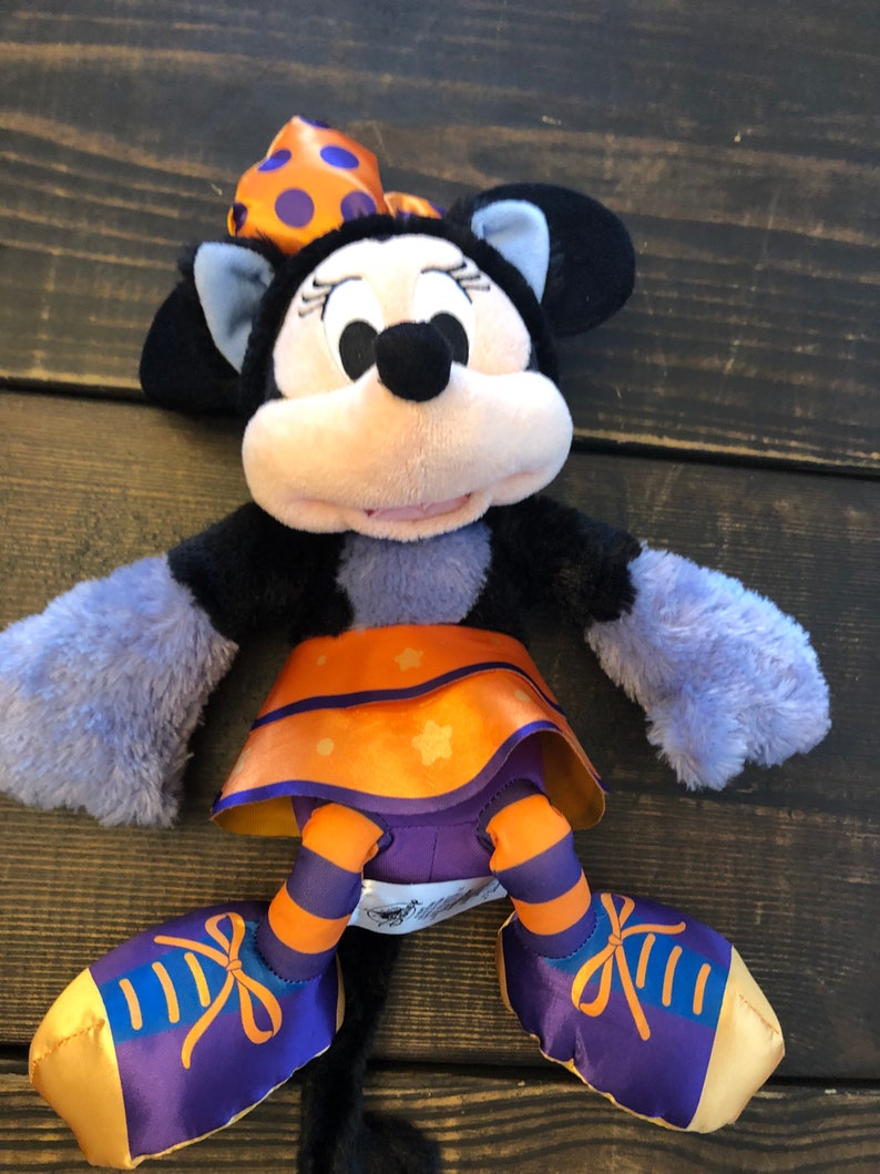 May include: A plush Minnie Mouse doll in a Halloween costume. The doll has a black head and ears, a light pink face, and a bow with orange polka dots. She wears a purple and orange skirt and shoes with orange stripes and bows.