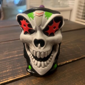 May include: A skull-shaped mug with a white face, black accents, and red starburst eye designs. The mug has a wide, toothy grin and a purple knob on the lid. Green accents are visible on the top and bottom of the mug.