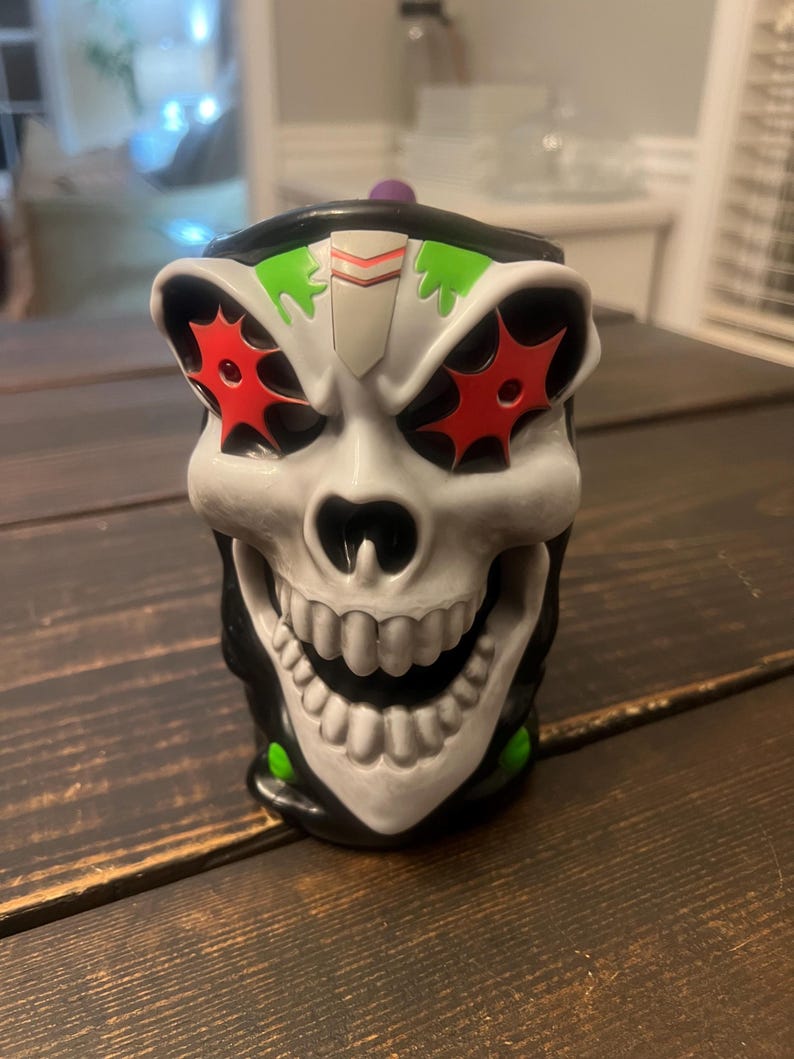May include: A skull-shaped mug with a white face, black accents, and red starburst eye designs. The mug has a wide, toothy grin and a purple knob on the lid. Green accents are visible on the top and bottom of the mug.