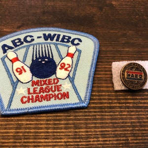 May include: A blue and red embroidered patch with the text "ABC-WIBC Mixed League Champion" and a bowling pin graphic. A small gold pin with the text "League Champion ABC 91-92" is also pictured.
