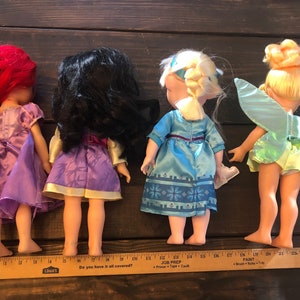 May include: Four Disney princess dolls, Ariel, Mulan, Elsa, and Tinkerbell, standing in a row. The dolls are all wearing their signature dresses and have long hair. The dolls are about 12 inches tall.