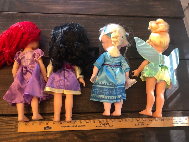 May include: Four Disney princess dolls, Ariel, Mulan, Elsa, and Tinkerbell, standing in a row. The dolls are all wearing their signature dresses and have long hair. The dolls are about 12 inches tall.