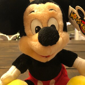 May include: A plush Mickey Mouse toy with black fur, large white eyes, and a red and white polka dot shorts. The toy has a yellow tag that says "Mouseketeers".