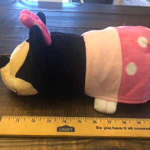May include: A plush toy shaped like a cartoon character with black fur, pink ears, and a pink and white polka dot dress. The toy is lying on its side with its head resting on the surface.