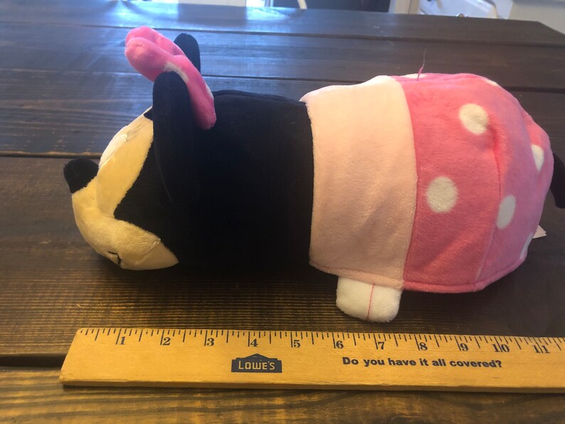 May include: A plush toy shaped like a cartoon character with black fur, pink ears, and a pink and white polka dot dress. The toy is lying on its side with its head resting on the surface.