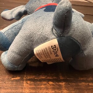 Op de afbeelding: Een lichtblauw pluche speelgoed van het personage Stitch uit Disney's Lilo & Stitch. Het speelgoed ligt op een houten oppervlak. Op het label van het speelgoed staat "Stitch" en "&copy;Disney".