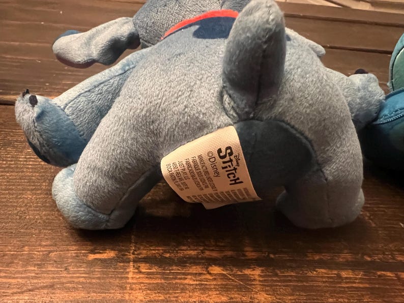 Op de afbeelding: Een lichtblauw pluche speelgoed van het personage Stitch uit Disney's Lilo & Stitch. Het speelgoed ligt op een houten oppervlak. Op het label van het speelgoed staat "Stitch" en "&copy;Disney".