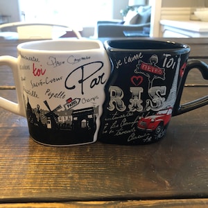 May include: Two white and black ceramic mugs with heart-shaped handles. The mugs are joined together and feature Parisian landmarks and text in French. The text on the white mug reads "Paris" and "toi". The text on the black mug reads "Je t'aime" and "Paris".