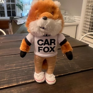 May include: A plush fox toy with orange and white fur, wearing a white t-shirt with "CAR FOX" printed in black, tan trousers, and white trainers. The fox has black paws and a black nose. The toy is standing on a dark wooden table.