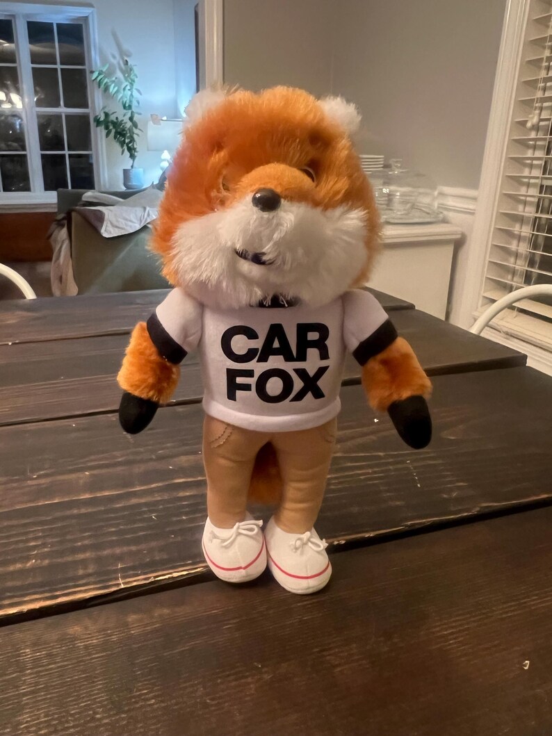 May include: A plush fox toy with orange and white fur, wearing a white t-shirt with "CAR FOX" printed in black, tan trousers, and white trainers. The fox has black paws and a black nose. The toy is standing on a dark wooden table.