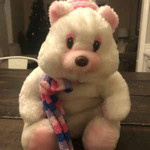 May include: White plush bear wearing a pink and blue striped hat and a pink, blue, and purple striped scarf. The bear has pink cheeks and a brown nose.