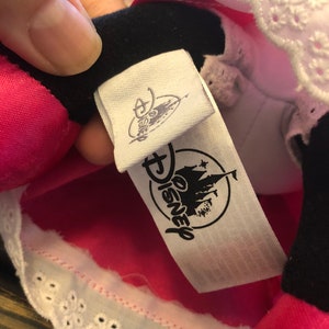 May include: A white tag with black text that reads "Disney" and features a black and white illustration of Cinderella's castle. The tag is attached to a pink and white garment with white lace trim.