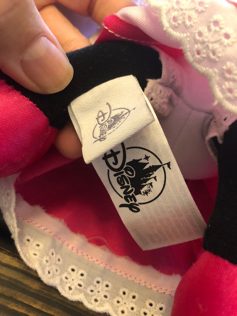May include: A white tag with black text that reads "Disney" and features a black and white illustration of Cinderella's castle. The tag is attached to a pink and white garment with white lace trim.