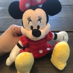Op de afbeelding: Een pluche speelgoed van Minnie Mouse die een rode jurk draagt met witte stippen en een rode strik met witte stippen. Ze heeft zwarte oren, een zwarte neus en gele schoenen.