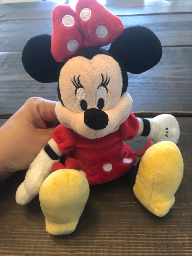 Op de afbeelding: Een pluche speelgoed van Minnie Mouse die een rode jurk draagt met witte stippen en een rode strik met witte stippen. Ze heeft zwarte oren, een zwarte neus en gele schoenen.