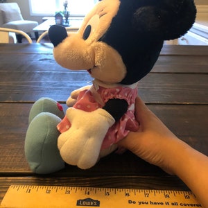 May include: A plush Minnie Mouse toy wearing a pink dress with white polka dots and a pink bow. The toy is sitting on a wooden surface with a ruler next to it. The ruler is marked with inches and has the words "Do you have it all covered" printed on it.