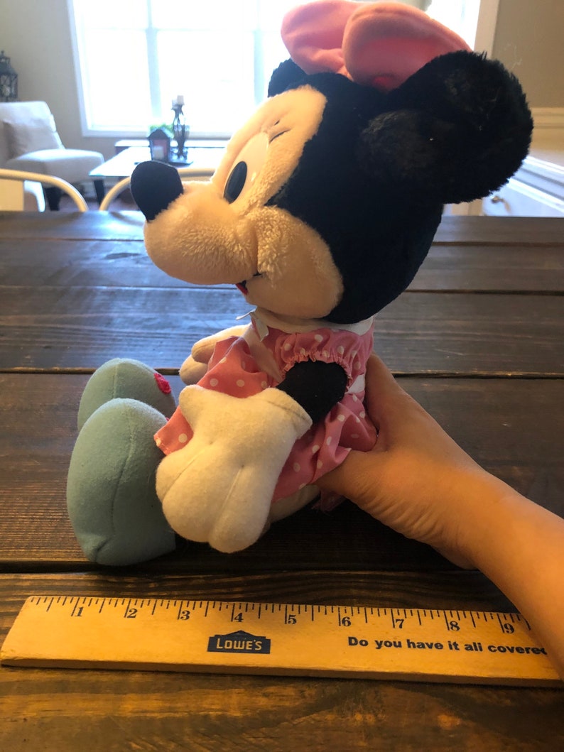 May include: A plush Minnie Mouse toy wearing a pink dress with white polka dots and a pink bow. The toy is sitting on a wooden surface with a ruler next to it. The ruler is marked with inches and has the words "Do you have it all covered" printed on it.