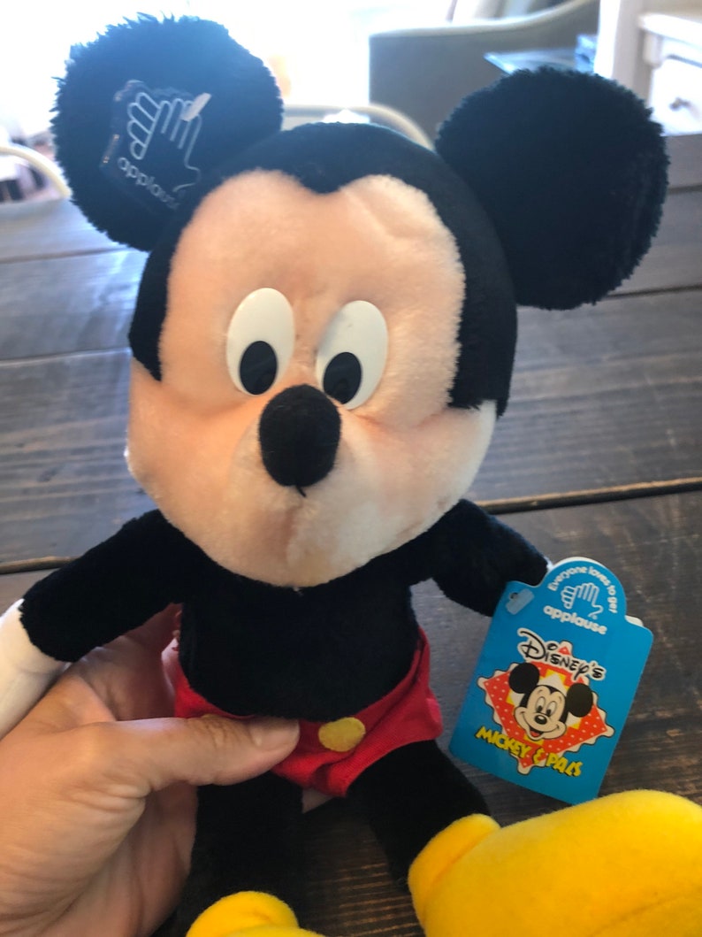 May include: A plush Mickey Mouse toy wearing red shorts with yellow buttons. The toy has a blue tag that says "Disney's Mickey & Pals" with a picture of Mickey Mouse on it.