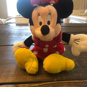 May include: A plush toy of Minnie Mouse wearing a red dress with white polka dots and a red bow with white polka dots. She has black ears and a black nose. Her feet are yellow.