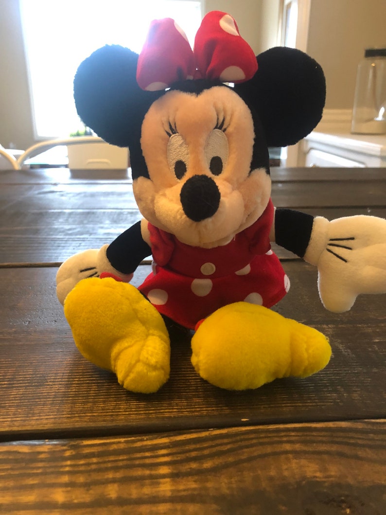 May include: A plush toy of Minnie Mouse wearing a red dress with white polka dots and a red bow with white polka dots. She has black ears and a black nose. Her feet are yellow.