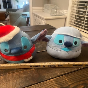 May include: Two plush Stitch toys, one in a Santa hat and scarf, the other in a winter outfit. Both are blue with large eyes and ears. A wooden ruler with black text lies in front of them.