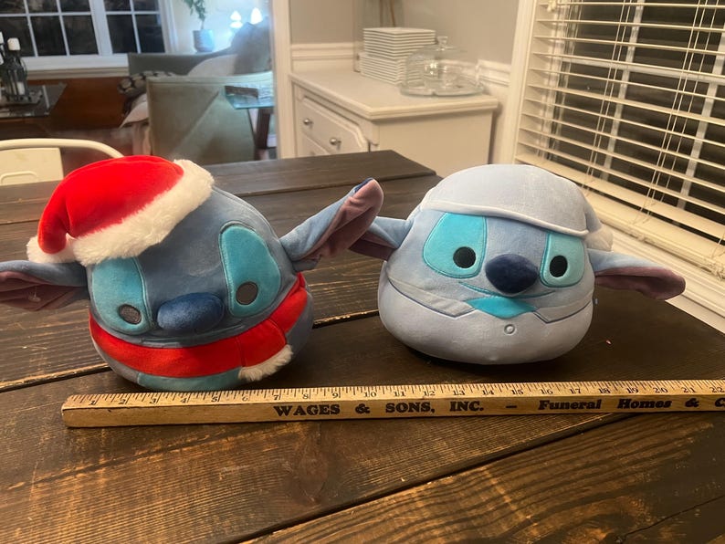May include: Two plush Stitch toys, one in a Santa hat and scarf, the other in a winter outfit. Both are blue with large eyes and ears. A wooden ruler with black text lies in front of them.