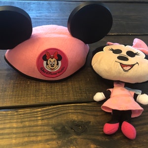 May include: Pink Minnie Mouse ears hat with a black bow and a red and white Minnie Mouse face patch that says "Walt Disney World". A small pink Minnie Mouse plush toy is also pictured.