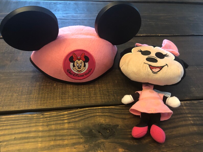 May include: Pink Minnie Mouse ears hat with a black bow and a red and white Minnie Mouse face patch that says "Walt Disney World". A small pink Minnie Mouse plush toy is also pictured.