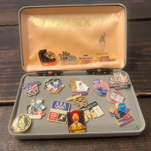 May include: A collection of vintage pins and badges, including Olympic and corporate logos, displayed in an open gray box. The pins feature various colors and designs, with text such as "BELLSOUTH" and "Atlanta 1996".