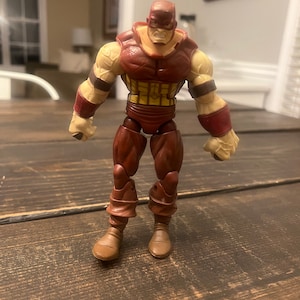 May include: A brown and red action figure of a superhero with a muscular build. The figure has a yellow and brown chest piece and brown boots.
