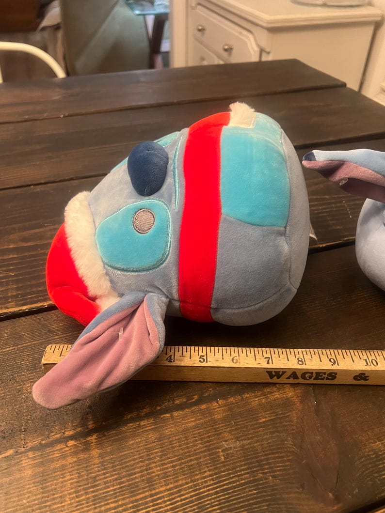 May include: A plush toy in the shape of a cartoon character's head, featuring blue, red, and white colors. The toy has large ears and a round head. A wooden ruler is placed next to the toy.