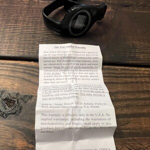May include: Black Armor All watch with a black band, resting on a wooden surface. The watch has a circular face with a digital display. A warranty document is next to the watch.