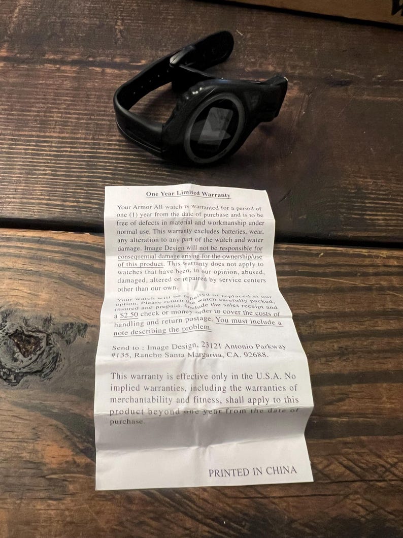 May include: Black Armor All watch with a black band, resting on a wooden surface. The watch has a circular face with a digital display. A warranty document is next to the watch.