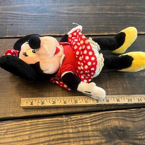 May include: A plush Minnie Mouse toy with black ears, a red dress with white polka dots, and yellow shoes. The toy is lying on a wooden surface with a ruler for scale.