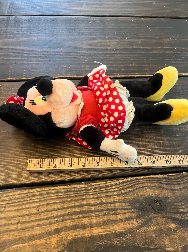 May include: A plush Minnie Mouse toy with black ears, a red dress with white polka dots, and yellow shoes. The toy is lying on a wooden surface with a ruler for scale.