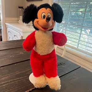 May include: A vintage Mickey Mouse plush toy. The plush has a black head with large ears, a red body, and white accents. The toy is standing on a white base and is positioned on a dark wooden table.