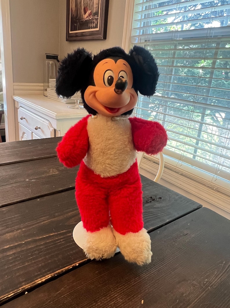 May include: A vintage Mickey Mouse plush toy. The plush has a black head with large ears, a red body, and white accents. The toy is standing on a white base and is positioned on a dark wooden table.