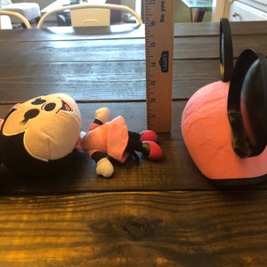 May include: A pink and black plush Minnie Mouse toy with a pink dress and black shoes. The toy is lying on a wooden table. There is a pink and black Minnie Mouse headband next to the toy.