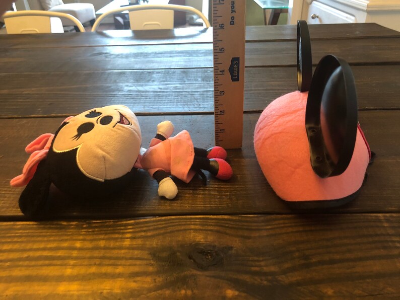 May include: A pink and black plush Minnie Mouse toy with a pink dress and black shoes. The toy is lying on a wooden table. There is a pink and black Minnie Mouse headband next to the toy.