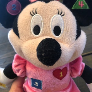 May include: Pink plush Minnie Mouse toy with embroidered eyes and a black nose. She is wearing a pink dress with a red heart with the number 1 on it, a blue square with the number 3 on it, and a green triangle with the number 4 on it. Her left ear has a purple circle with the number 2 on it.