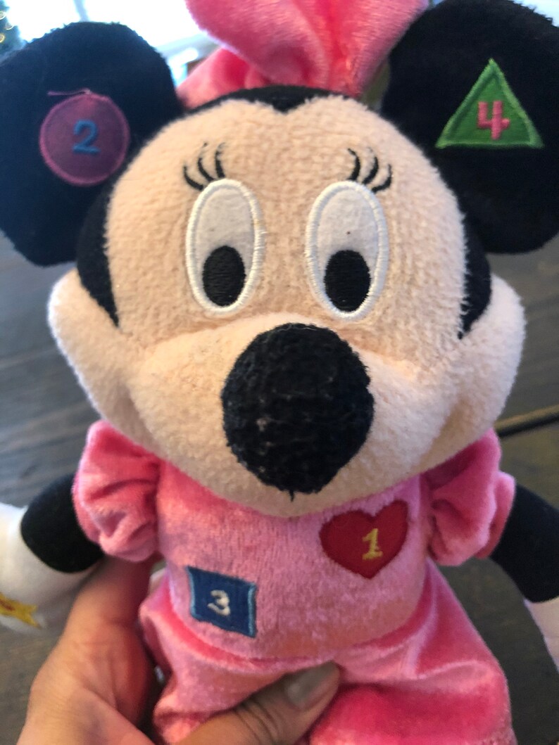 May include: Pink plush Minnie Mouse toy with embroidered eyes and a black nose. She is wearing a pink dress with a red heart with the number 1 on it, a blue square with the number 3 on it, and a green triangle with the number 4 on it. Her left ear has a purple circle with the number 2 on it.