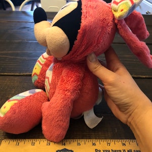 Op de afbeelding: Roze pluche Mickey Mouse verkleed als paashaas met een kleurrijke gestreepte strik. Het pluche speelgoed is ongeveer 15 cm hoog en heeft een wit label op zijn voet.