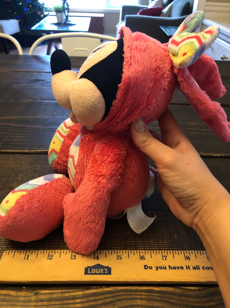 Op de afbeelding: Roze pluche Mickey Mouse verkleed als paashaas met een kleurrijke gestreepte strik. Het pluche speelgoed is ongeveer 15 cm hoog en heeft een wit label op zijn voet.