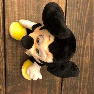 Puede incluir: Un juguete de peluche Mickey Mouse negro y amarillo con orejas grandes. El juguete tiene una cara sonriente con ojos marrones y una nariz negra.