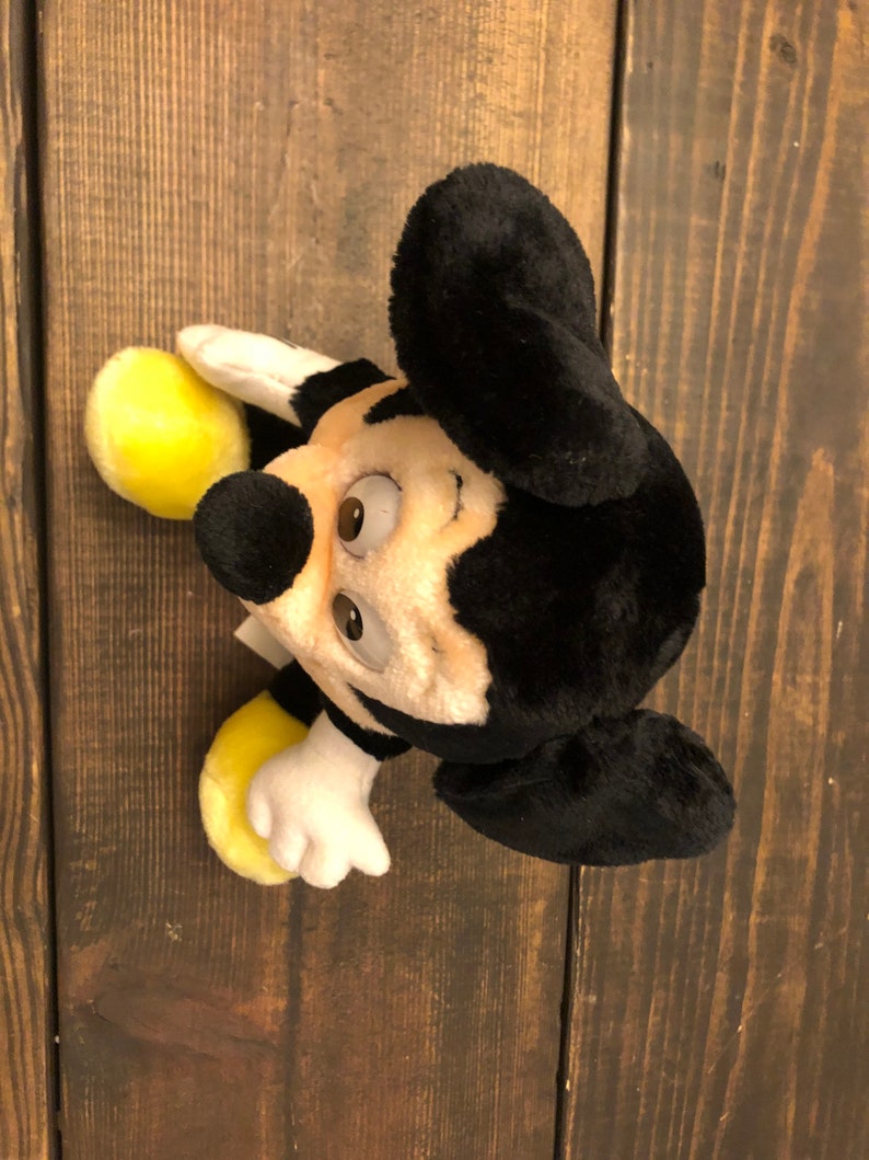 Puede incluir: Un juguete de peluche Mickey Mouse negro y amarillo con orejas grandes. El juguete tiene una cara sonriente con ojos marrones y una nariz negra.